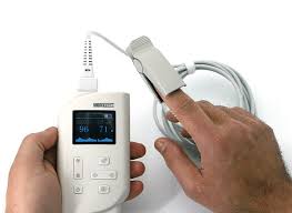 A handheld pulse oximeter monitor held by two hands with a finger clip probe on a finger, displaying SpO2 and heart rate.
