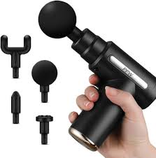 Handheld massage gun with multiple interchangeable attachments laid out beside it; black device with a hand gripping the handle.